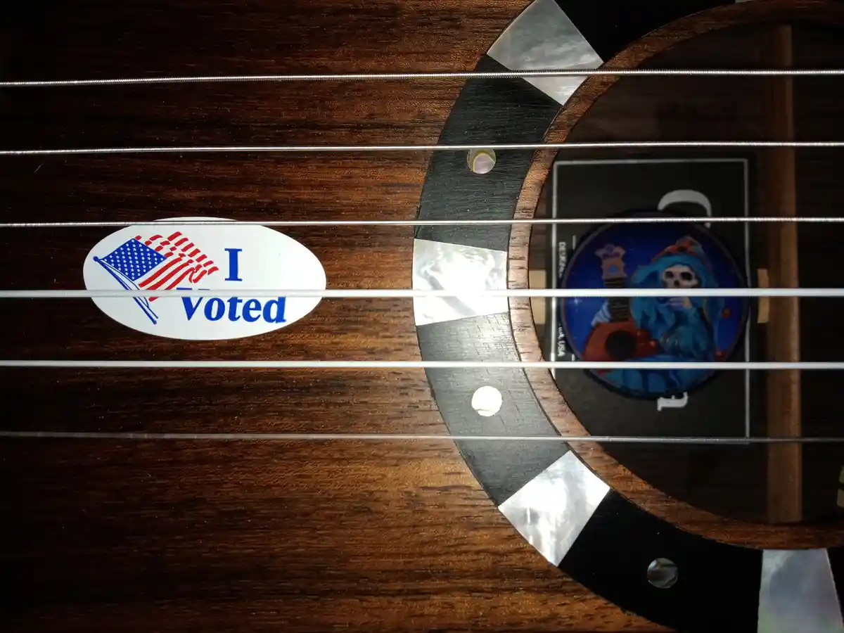 A close-up of a dark wood guitar body with six metal strings, showing an 'I Voted' sticker with an American flag design on…