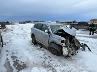 A silver SUV is severely damaged with its front end crushed and mangled, parked in a snowy, muddy lot. Two people stand in…