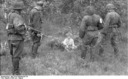 A black-and-white historical photograph shows five soldiers in World War II-era German uniforms standing in a grassy field…
