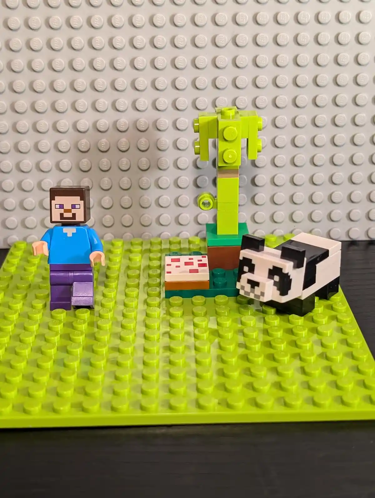 A Lego Minecraft Steve minifigure stands beside a Lego panda and a small tree on a green baseplate, with a red and white…