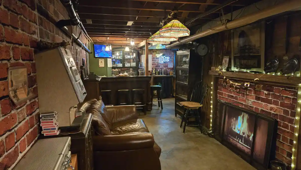 A cozy, dimly lit basement bar with exposed brick walls, a leather couch, a fireplace with a glowing fire, and a bar area…