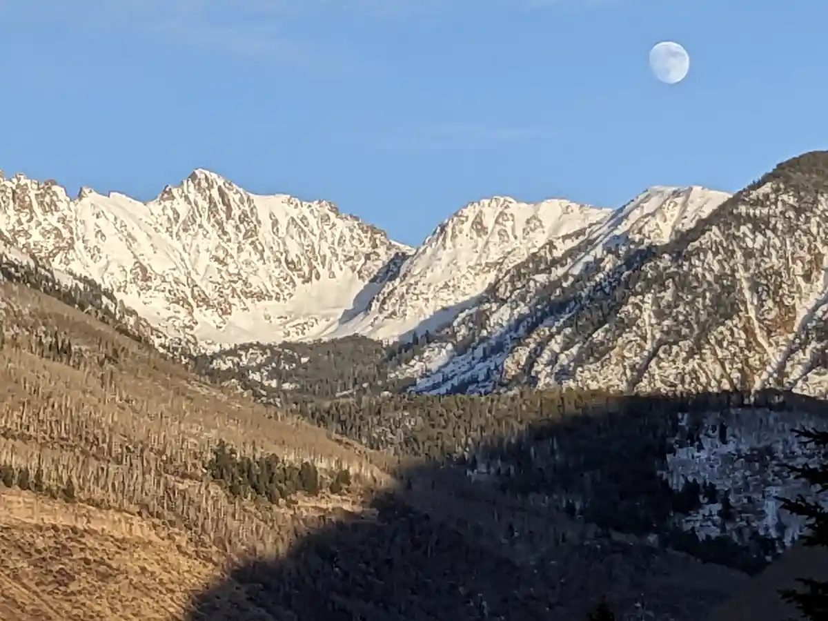 A scenic photo of snow-capped mountains under a clear blue sky with the moon visible in the upper right corner. The overlaid…