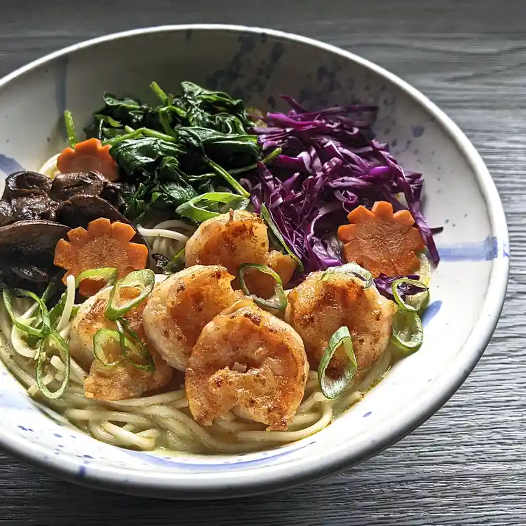 A close-up photo of a bowl of ramen with noodles, shrimp, spinach, purple cabbage, mushrooms, and flower-shaped carrot…