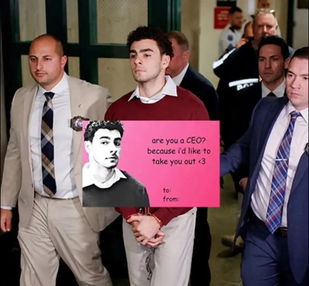 A man in a red sweater, handcuffed and flanked by law enforcement officers, holds a pink card with a black-and-white photo…