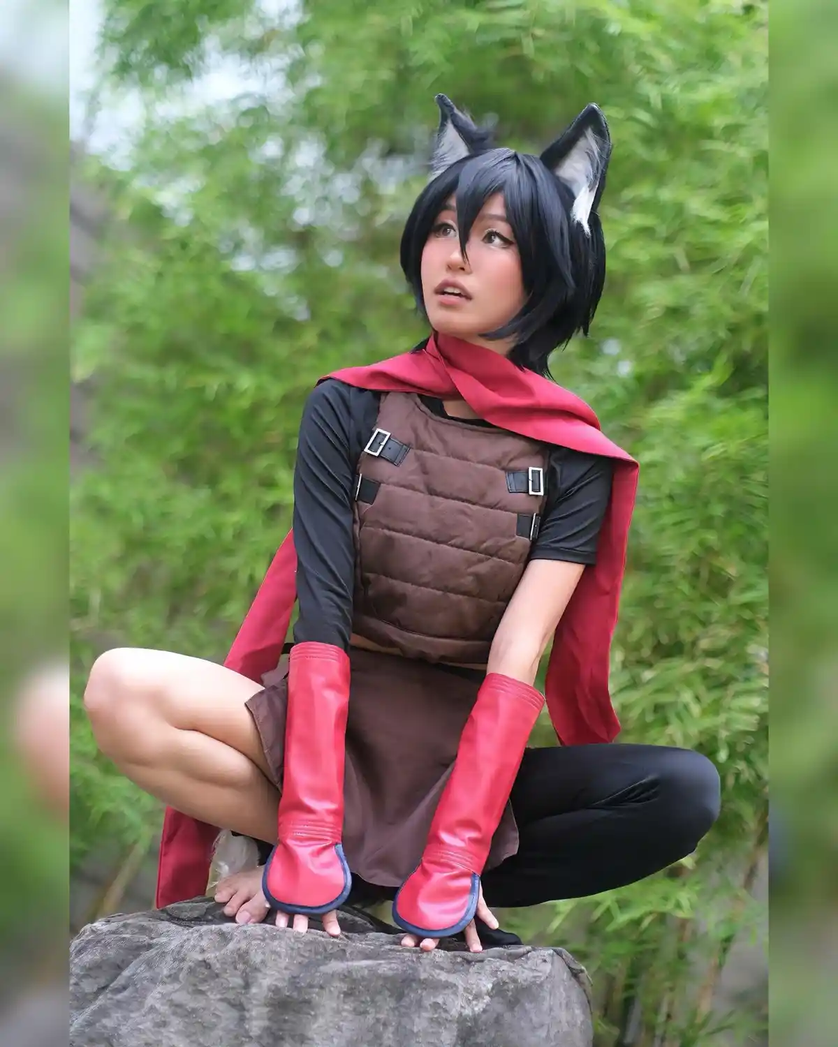 A person in a cosplay outfit with black wolf ears, a red cape, brown vest, and red gloves sits on a rock outdoors. The text…