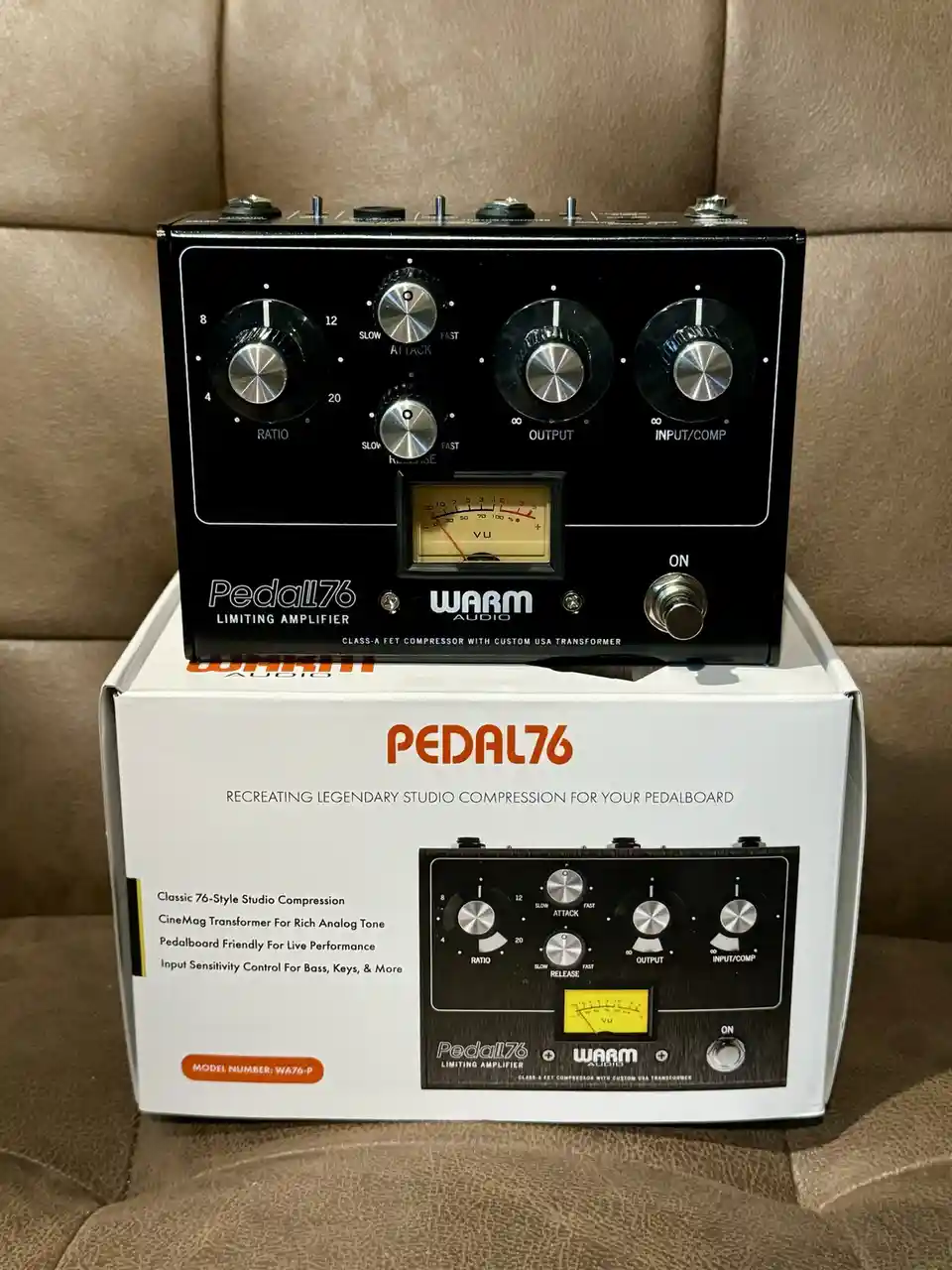 A black Warm Audio Pedal76 limiting amplifier sits atop its white retail box, both featuring identical control knobs labeled…