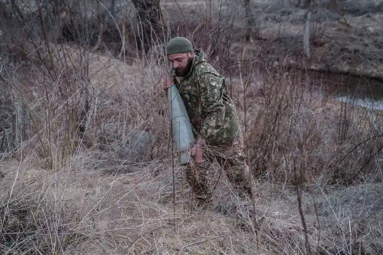 A man in military camouflage gear and a green beanie is crouching in a field of dry brush, holding a large, cylindrical…