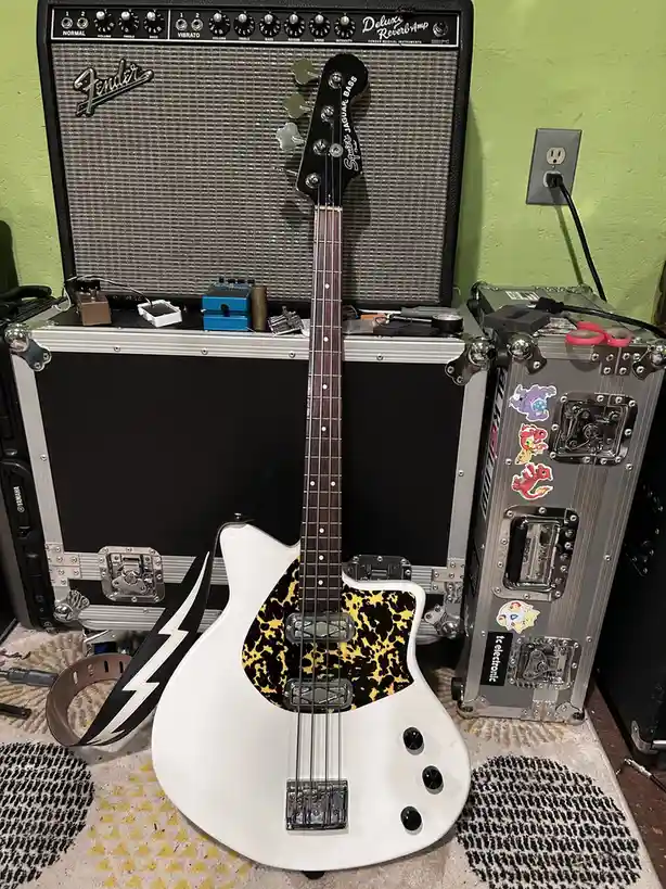 A white Fender Jaguar Bass guitar with a leopard-print pickguard stands upright on a patterned rug, leaning against a Fender…