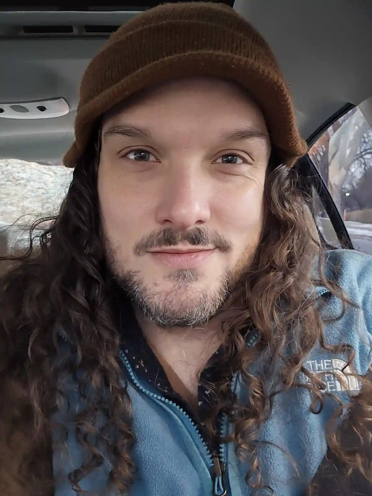 A selfie of a man with long curly brown hair and a brown beanie, wearing a blue fleece jacket, sitting inside a car. He has…