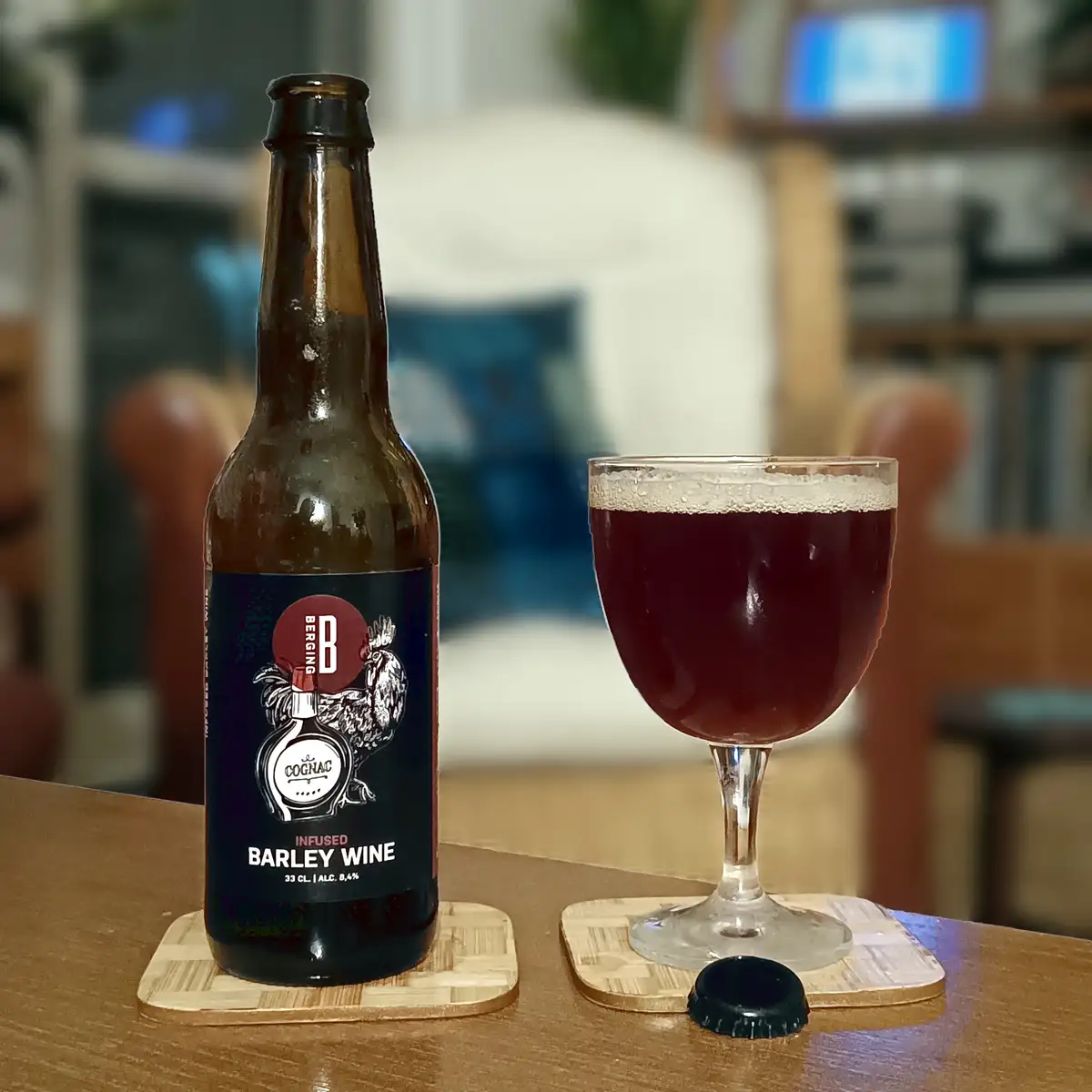 A brown bottle labeled 'Berging B Infused Barley Wine' sits next to a glass of dark red beverage on a wooden table, with the…
