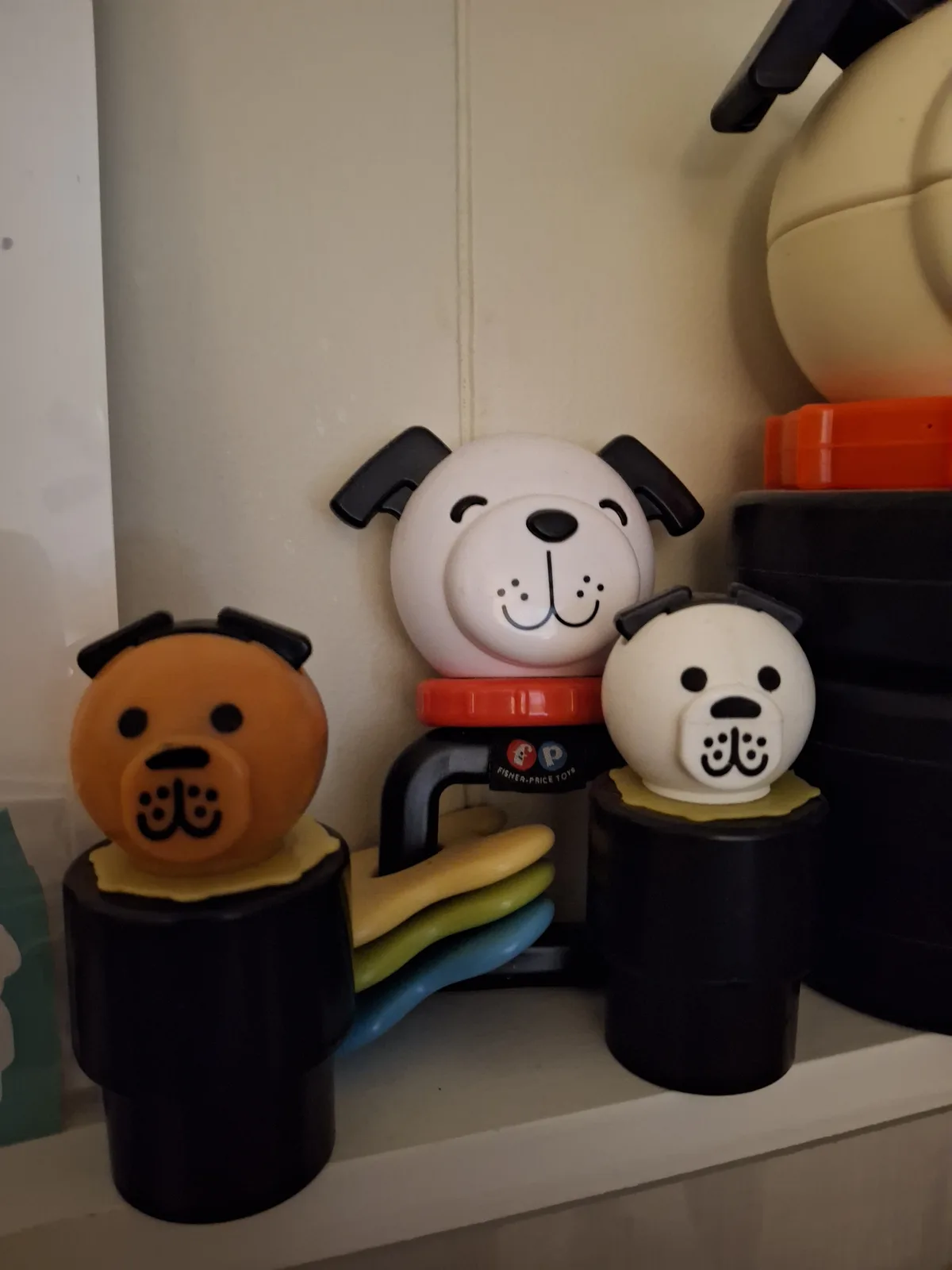 Three Fisher-Price dog-shaped baby toys sit on a shelf, two white and one orange, all with simple cartoon faces. The central…