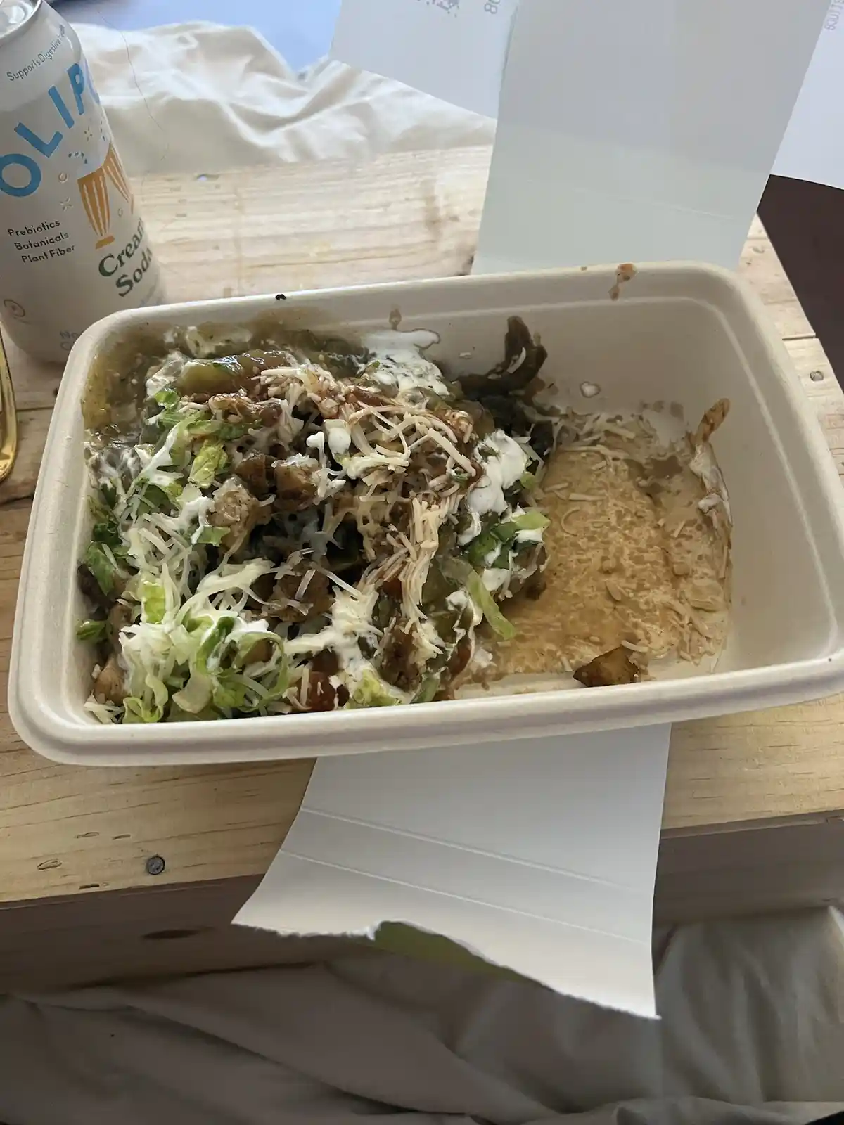 A partially eaten burrito bowl in a white takeout container sits on a wooden table, showing mostly toppings like shredded…