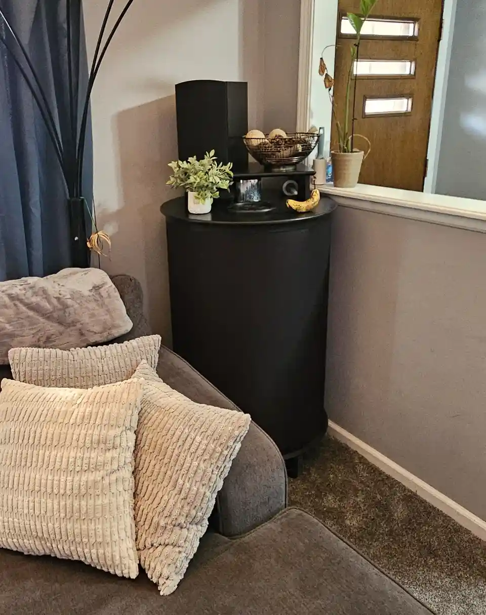A cozy living room corner with a gray couch adorned with textured beige pillows, next to a black cylindrical side table…