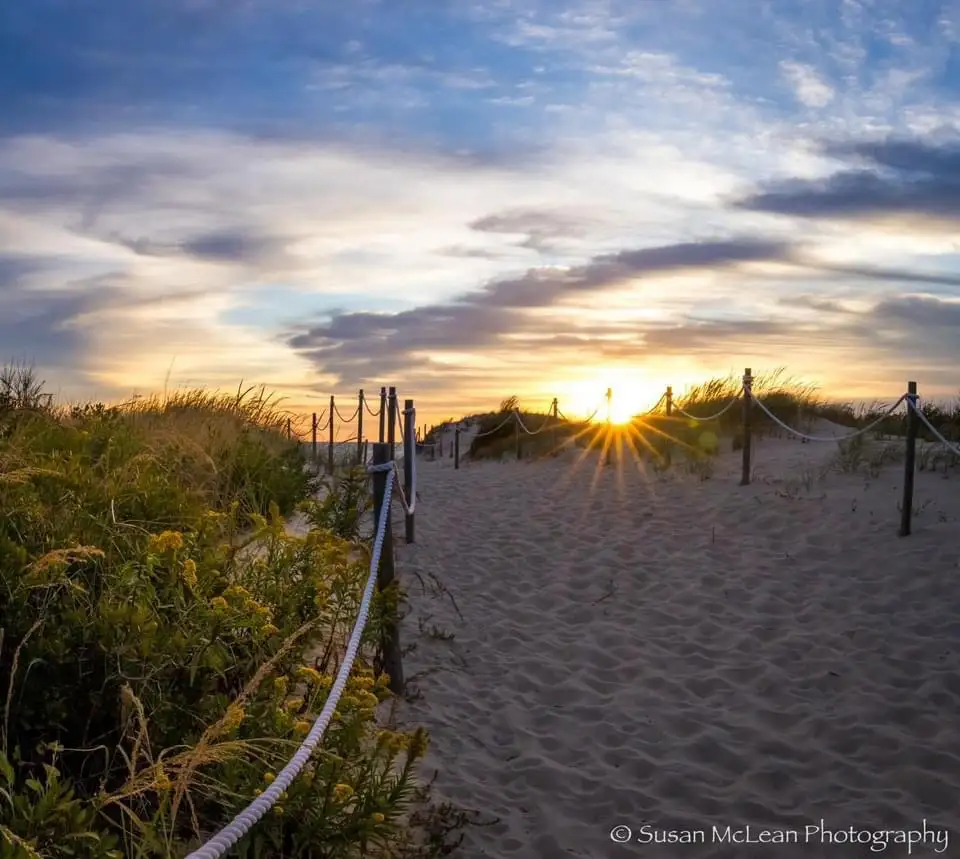 A serene beach scene at sunset with a sandy path flanked by wooden posts and rope, leading toward the sun. The sky is…