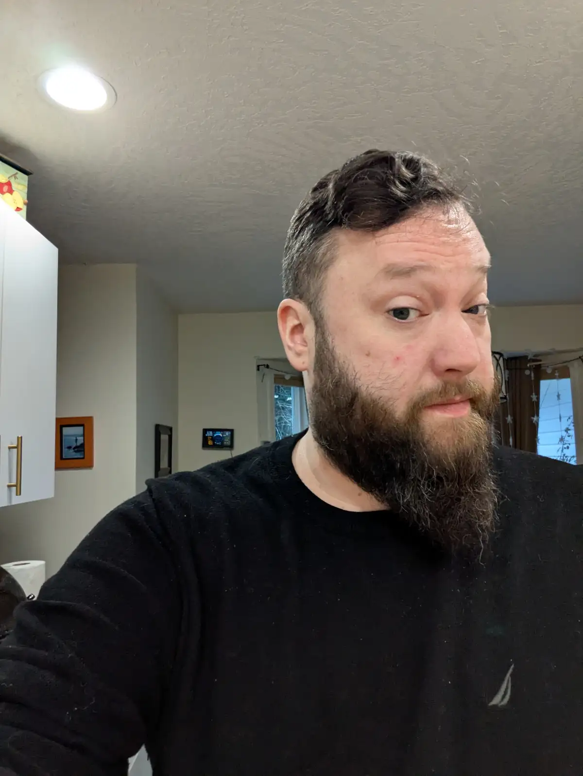 A man with a full beard and short dark hair, wearing a black long-sleeved shirt, looks directly at the camera with a…