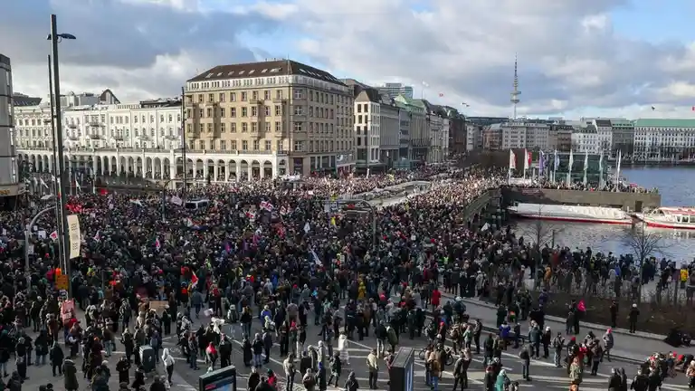 A wide-angle photograph shows a massive crowd of people gathered in a city square near a river, holding signs and banners…