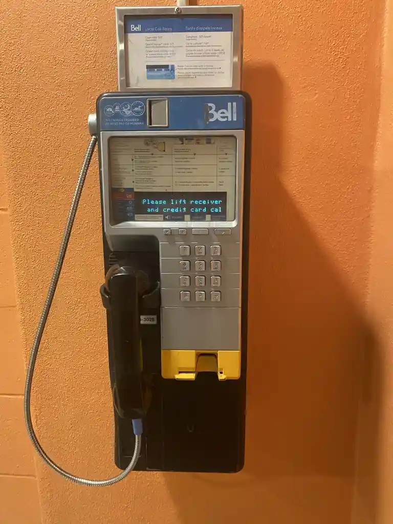 A Bell payphone mounted on an orange wall displays a glitched message on its screen that reads 'Please lift receiver and…