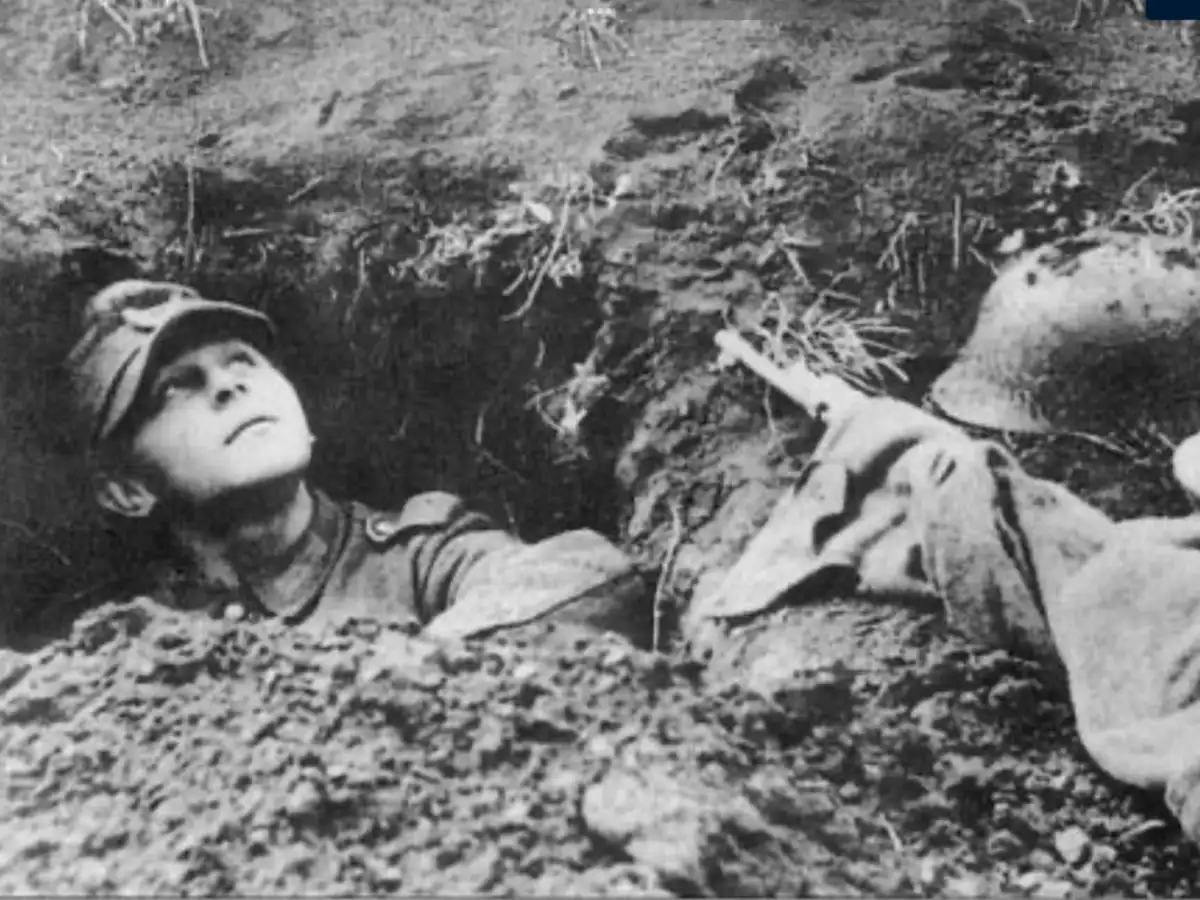 A black-and-white historical photo shows a young soldier lying in a muddy trench, looking upward with a calm expression…
