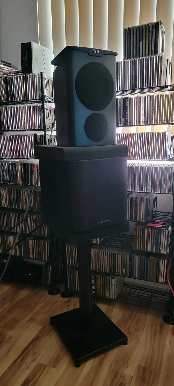 A tall black ELAC speaker sits atop a larger Bowers & Wilkins subwoofer, both mounted on a stand in a room filled with CD…