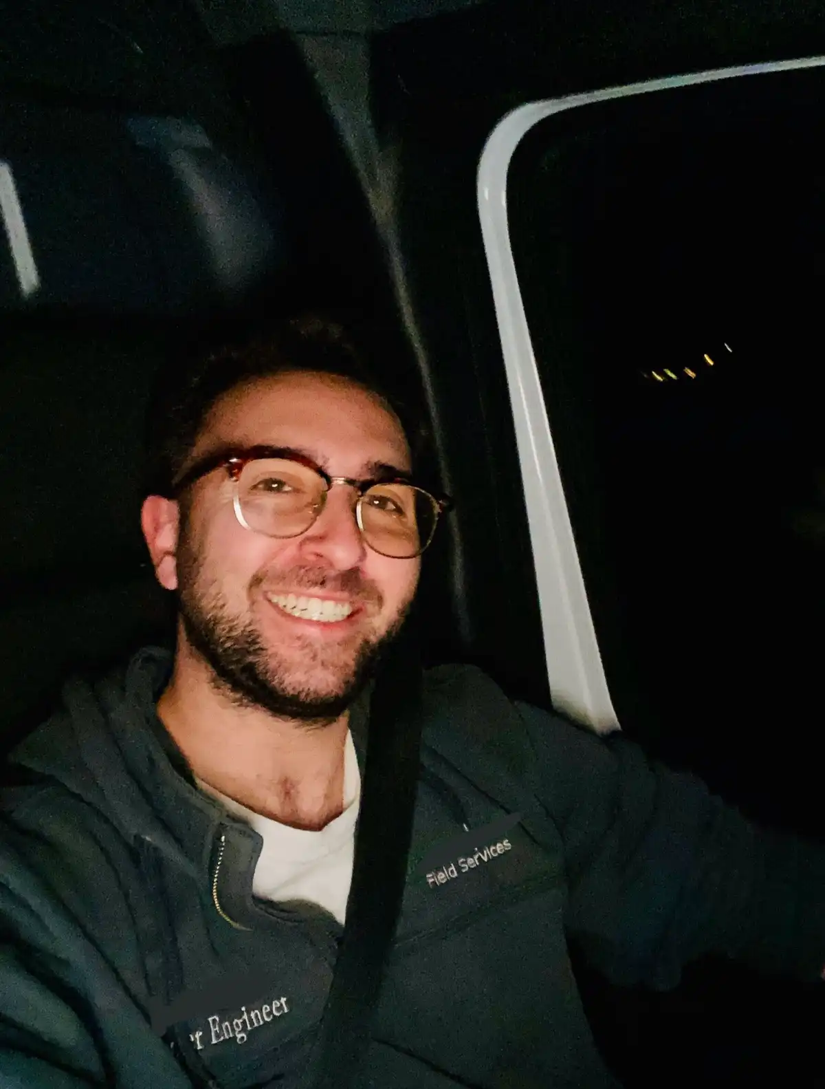 A smiling man with glasses and a beard sits in a car at night, wearing a dark jacket with 'Field Services' and 'Engineer'…