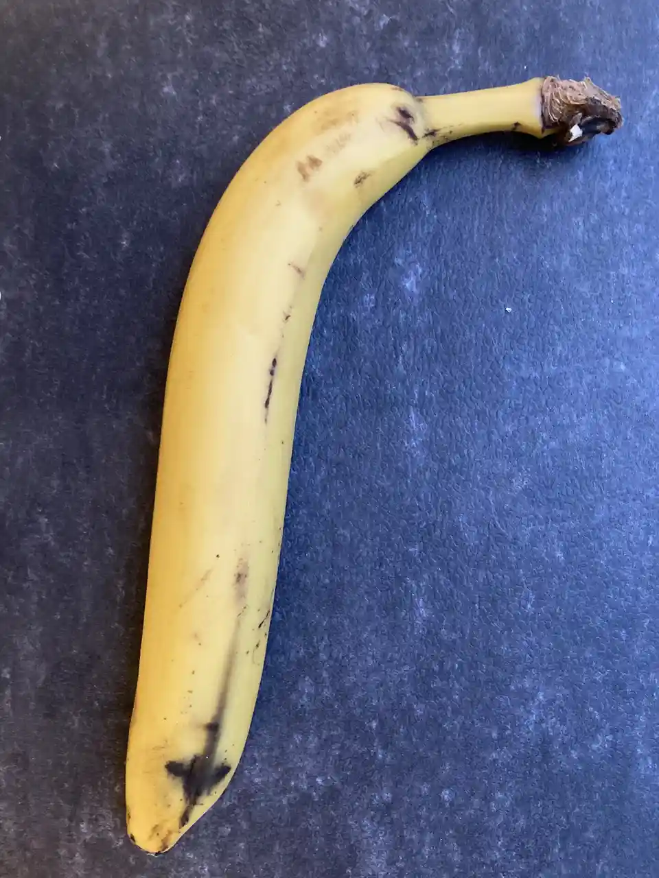 A single yellow banana with brown spots and a curved shape rests on a dark blue textured surface. The banana's peel shows…
