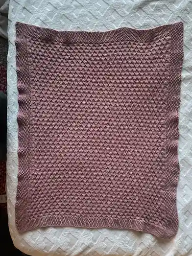 A dusty rose-colored knitted blanket with a textured, repeating diamond-like pattern lies flat on a white quilted surface…