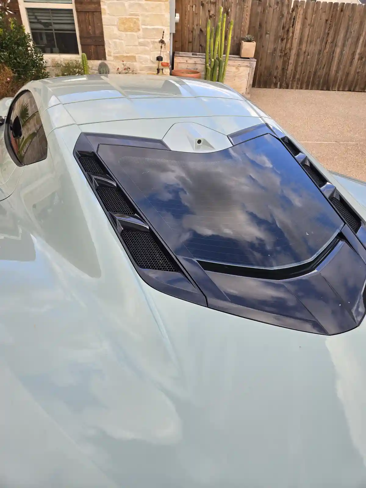 A high-angle shot of a light-colored sports car's hood, which has a large black panel with vents and a camera. The hood…