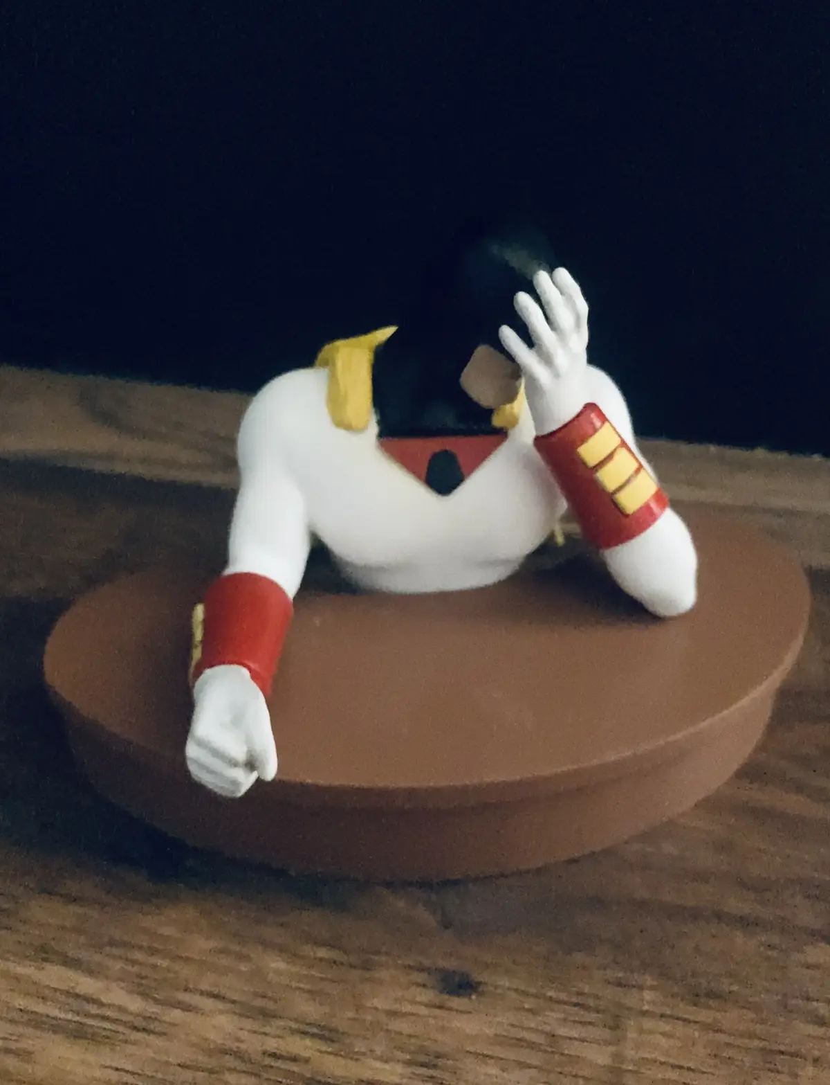 A small figurine of a superhero with a white suit, red and yellow accents, and black hair is posed slumped over a brown…