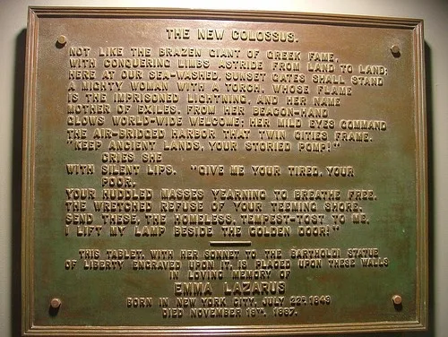 A close-up photograph of a bronze plaque with engraved text, showing the full poem 'The New Colossus' by Emma Lazarus, with…