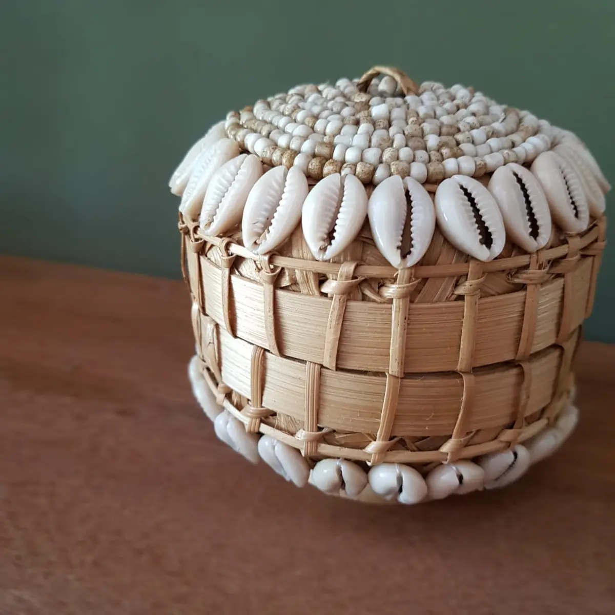 A close-up photo of a handcrafted, cylindrical woven basket with a lid, decorated with rows of white cowrie shells and small…
