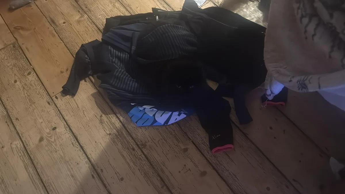 A black cat is nestled inside a pile of clothes on a wooden floor, looking up with wide eyes. The overlaid text reads 'i'm…