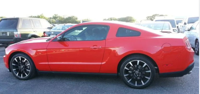 A bright red Ford Mustang coupe is parked in a lot, viewed from the side. The car has black multi-spoke wheels and a glossy…