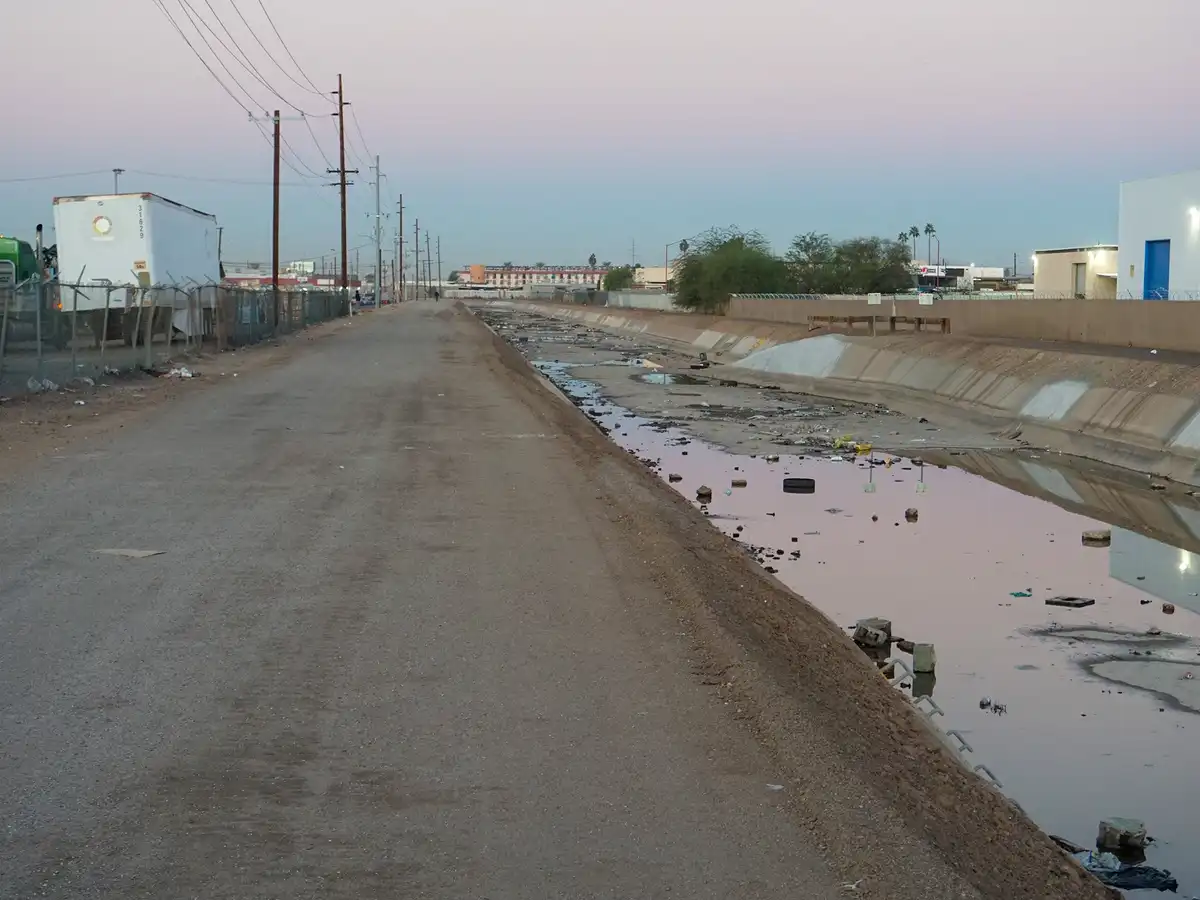 A long, straight, unpaved road runs parallel to a dirty, littered drainage canal filled with murky water and debris. On the…