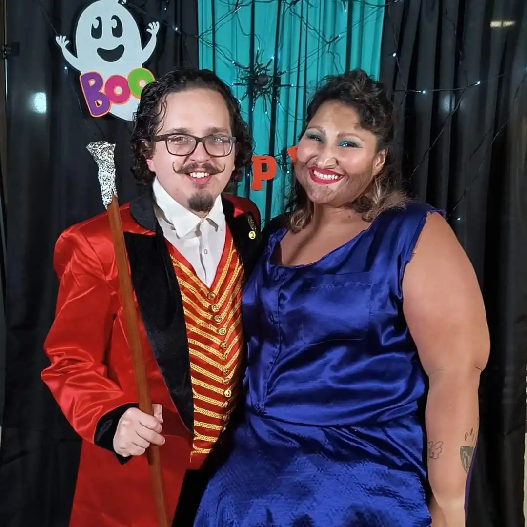 A man in a red and gold ringmaster costume with a mustache and glasses holds a cane, standing beside a woman in a blue satin…