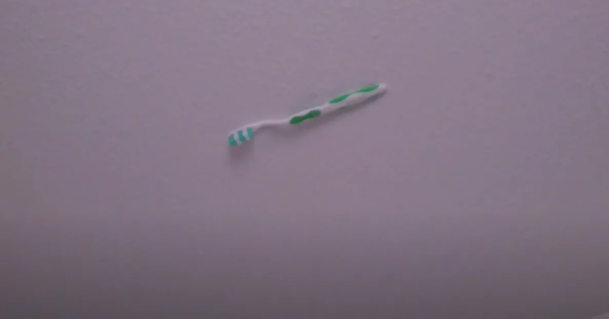 A blurry image shows a green and white toothbrush lying horizontally on a plain, textured, light-colored ceiling. The…