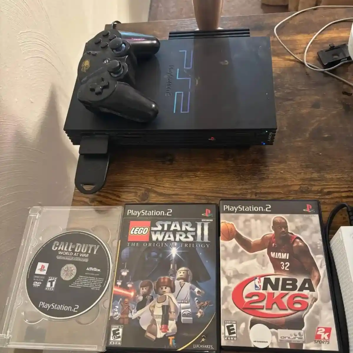 A black PlayStation 2 console sits on a wooden table with a black controller resting on top. In front of the console are…