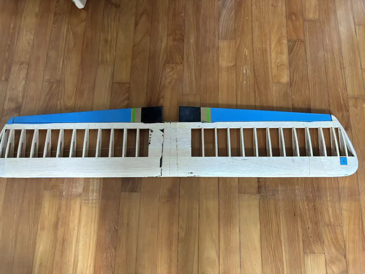A wooden model airplane fuselage lies on a hardwood floor, its wings partially painted blue with black and green tape…