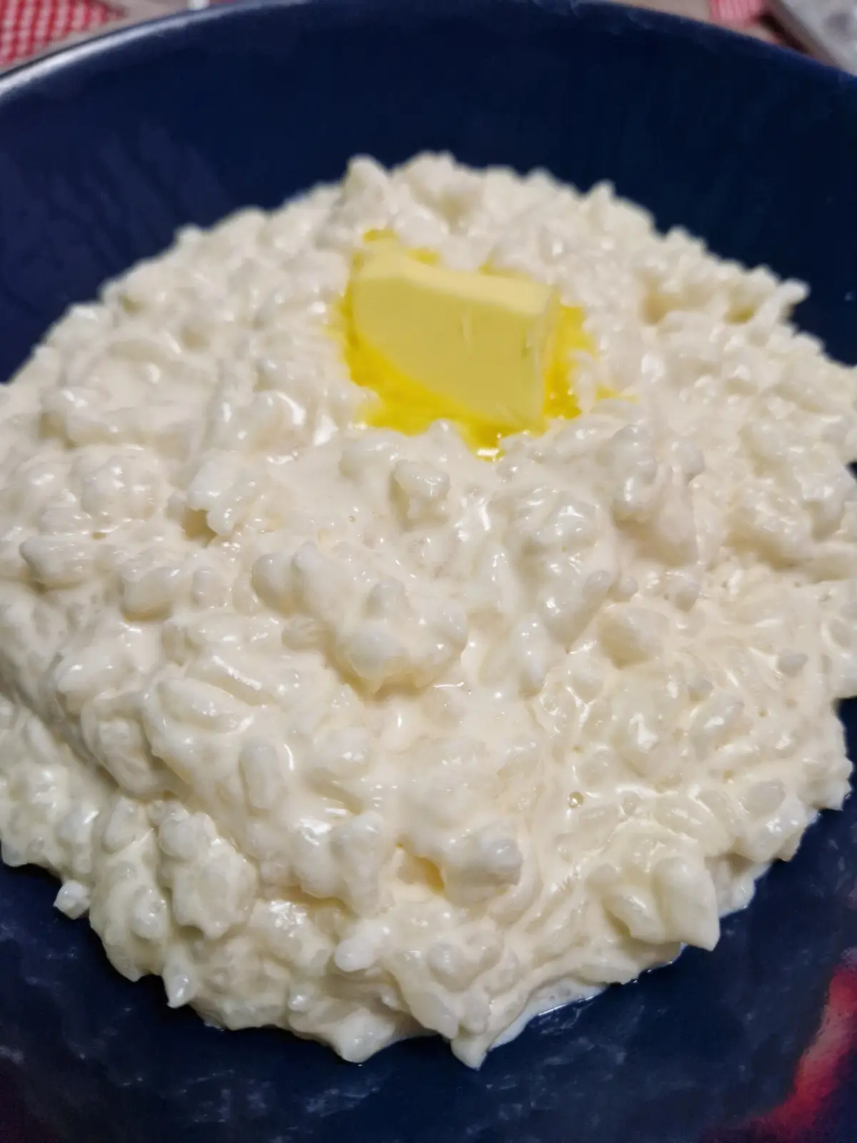 A close-up photo shows a creamy, lumpy white risotto in a dark blue bowl, with a single pat of yellow butter melting on top…