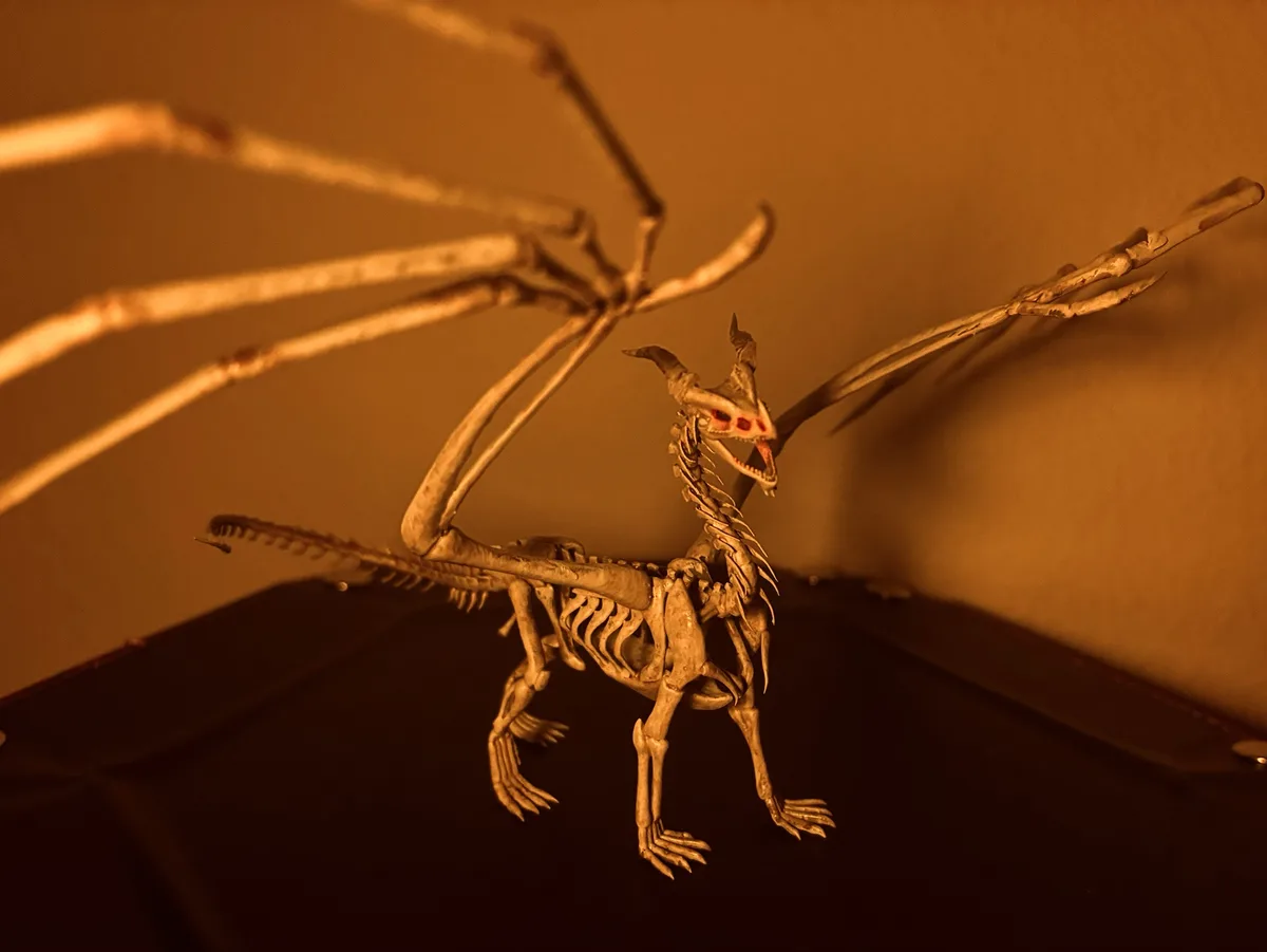 A skeletal dragon model with spread wings and glowing red eyes stands on a dark surface under warm, orange lighting. The…