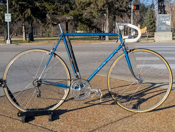A blue vintage road bicycle with white handlebar tape and yellow-rimmed wheels is parked on a gravel sidewalk next to a…