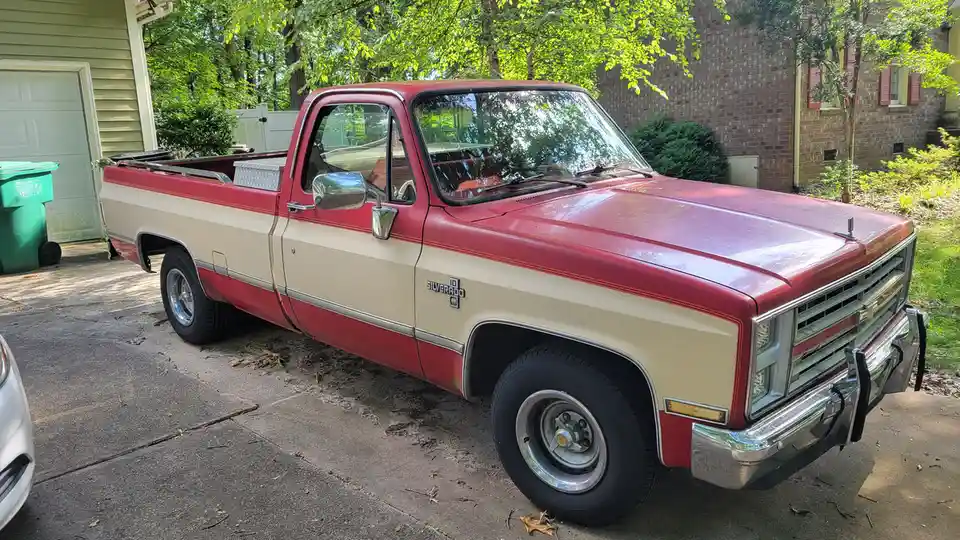 A red and beige 1980s Chevrolet Silverado pickup truck is parked in a driveway next to a house, with trees and a green…