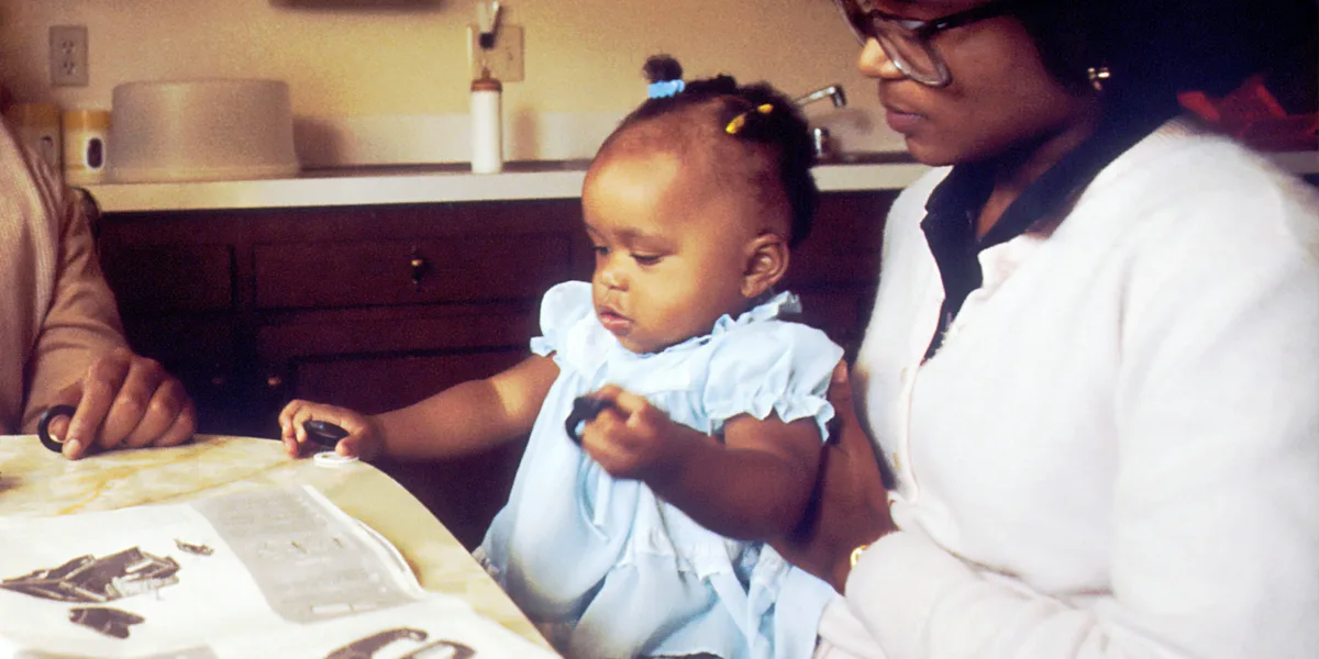 A vintage photo shows a young Black child in a light blue dress with pigtails, sitting at a table and looking down with a…