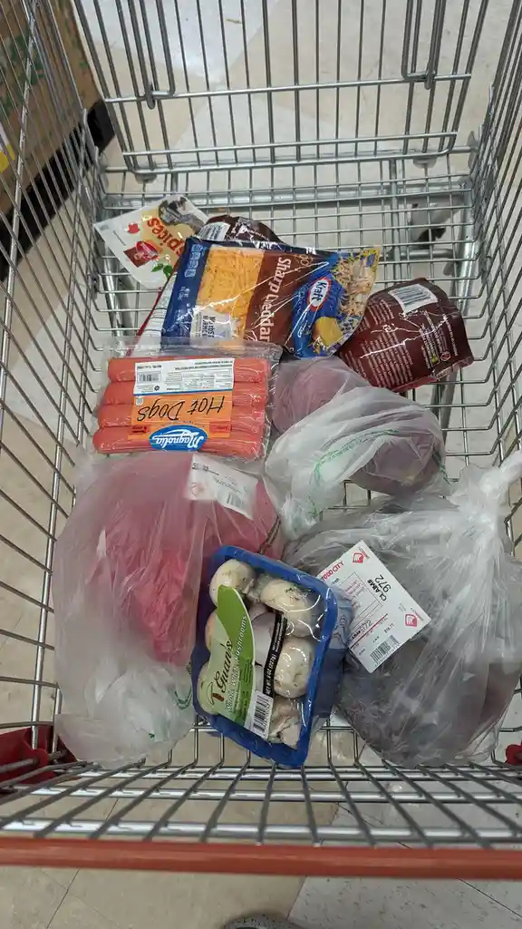 A shopping cart filled with groceries including a package of hot dogs with 'Hot Dogs' handwritten on it, a bag of whole…
