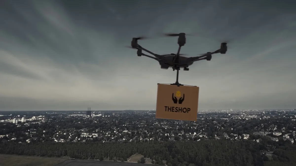 A drone hovers over a sprawling cityscape under a cloudy sky, carrying a cardboard box labeled 'THESHOP' with a logo of two…
