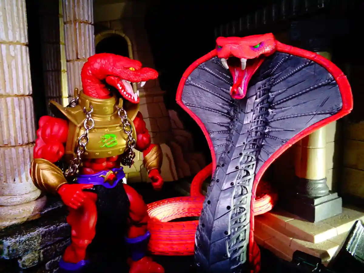 Two red snake-themed action figures are posed in a stone-floored, columned setting. The figure on the left, resembling a…