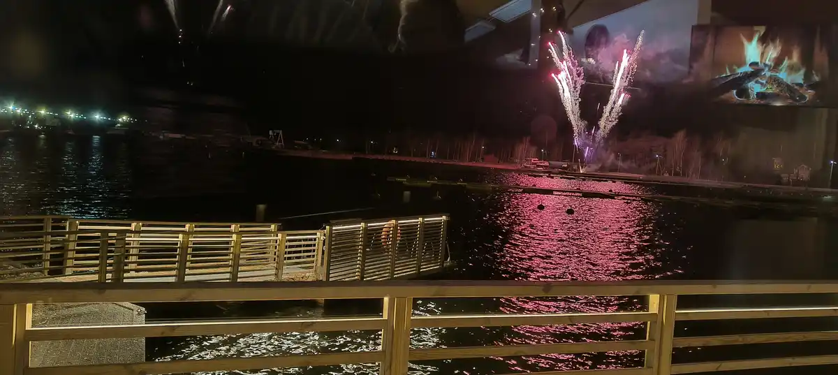 A nighttime photo shows colorful fireworks exploding over a body of water, with their reflections shimmering on the surface…