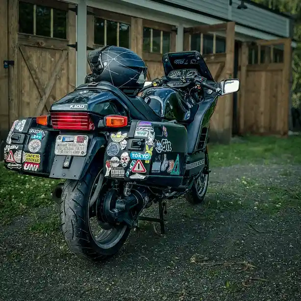 A black BMW K100 motorcycle is parked on gravel in front of a wooden garage, covered in numerous colorful stickers on its…