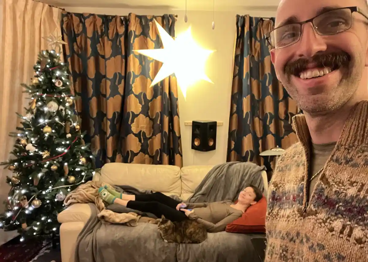 A man with glasses and a mustache smiles at the camera in a cozy living room with a decorated Christmas tree and a lit star…