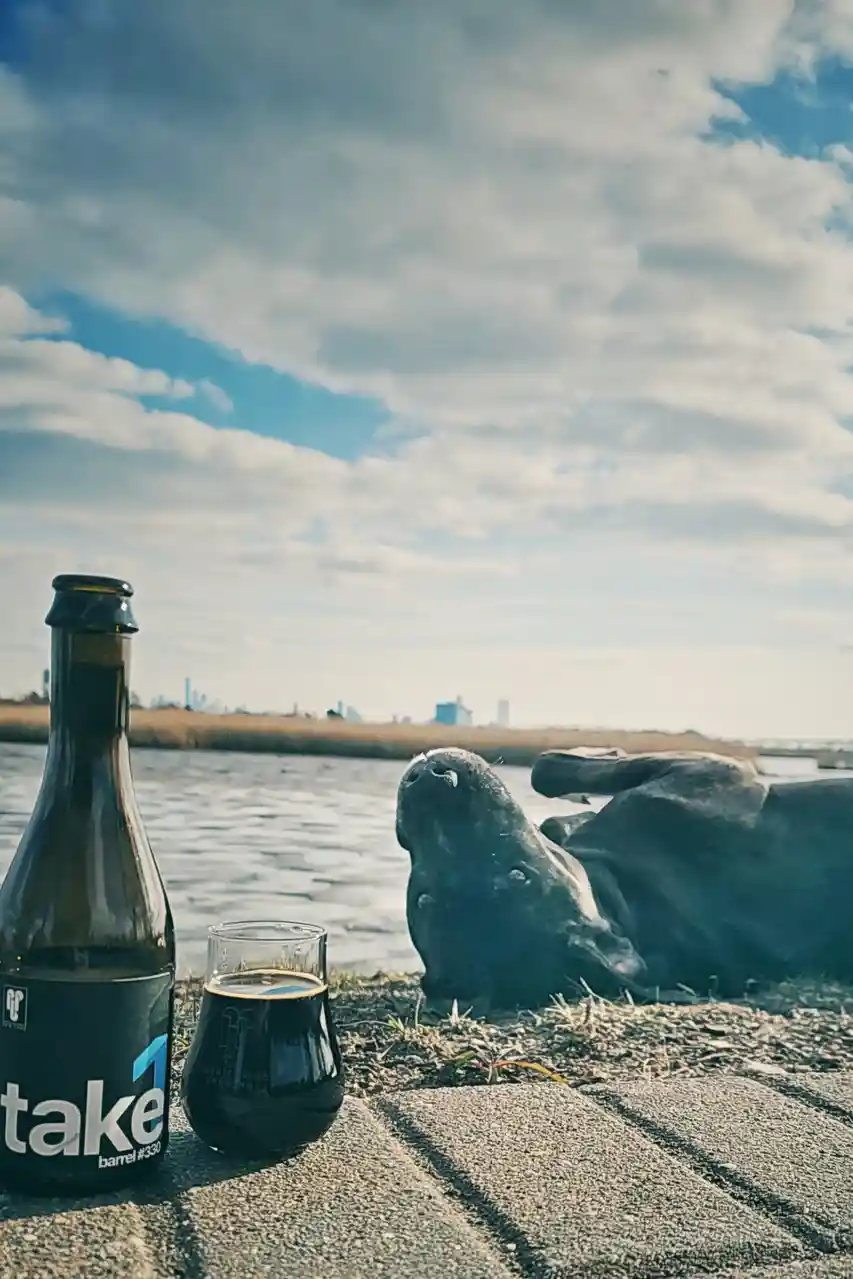 A dark beer bottle and glass sit on a paved surface near water, next to a large bronze sculpture of a dog lying on its back…