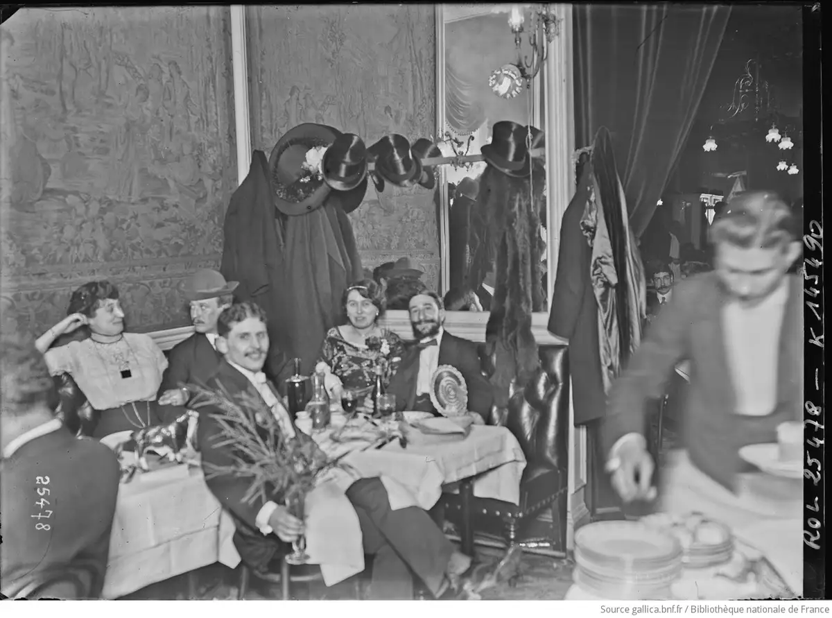 A vintage black-and-white photograph shows a group of elegantly dressed people seated at a restaurant table, smiling at the…